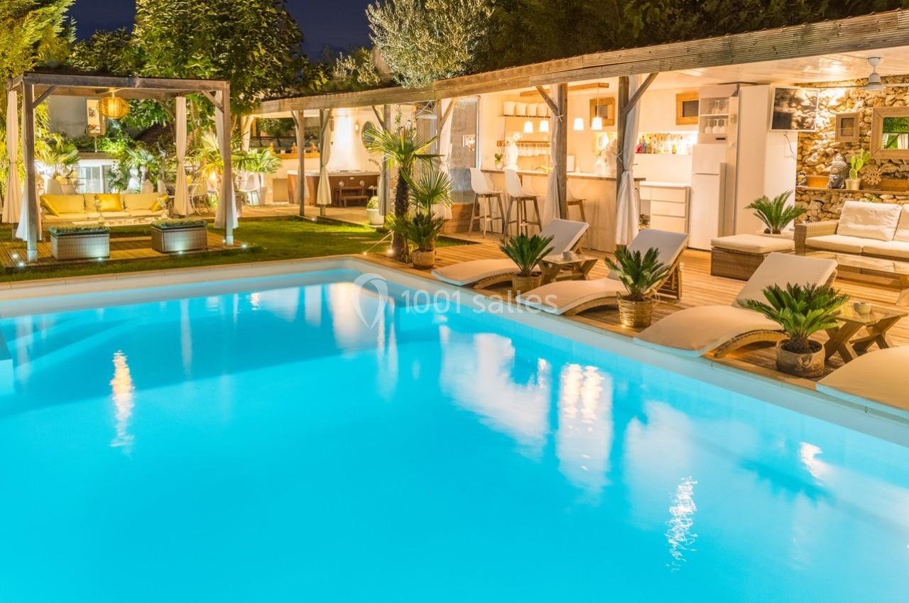 Terrasse aménagée avec piscine éclairée, transats, végétation et espace bar sous un éclairage chaleureux en soirée.