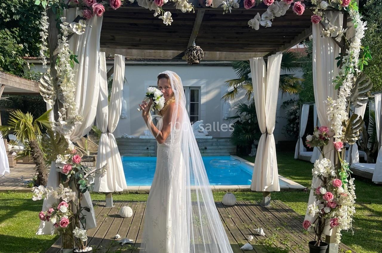 Une mariée en robe blanche pose sous une pergola décorée de fleurs, avec une piscine en arrière-plan.