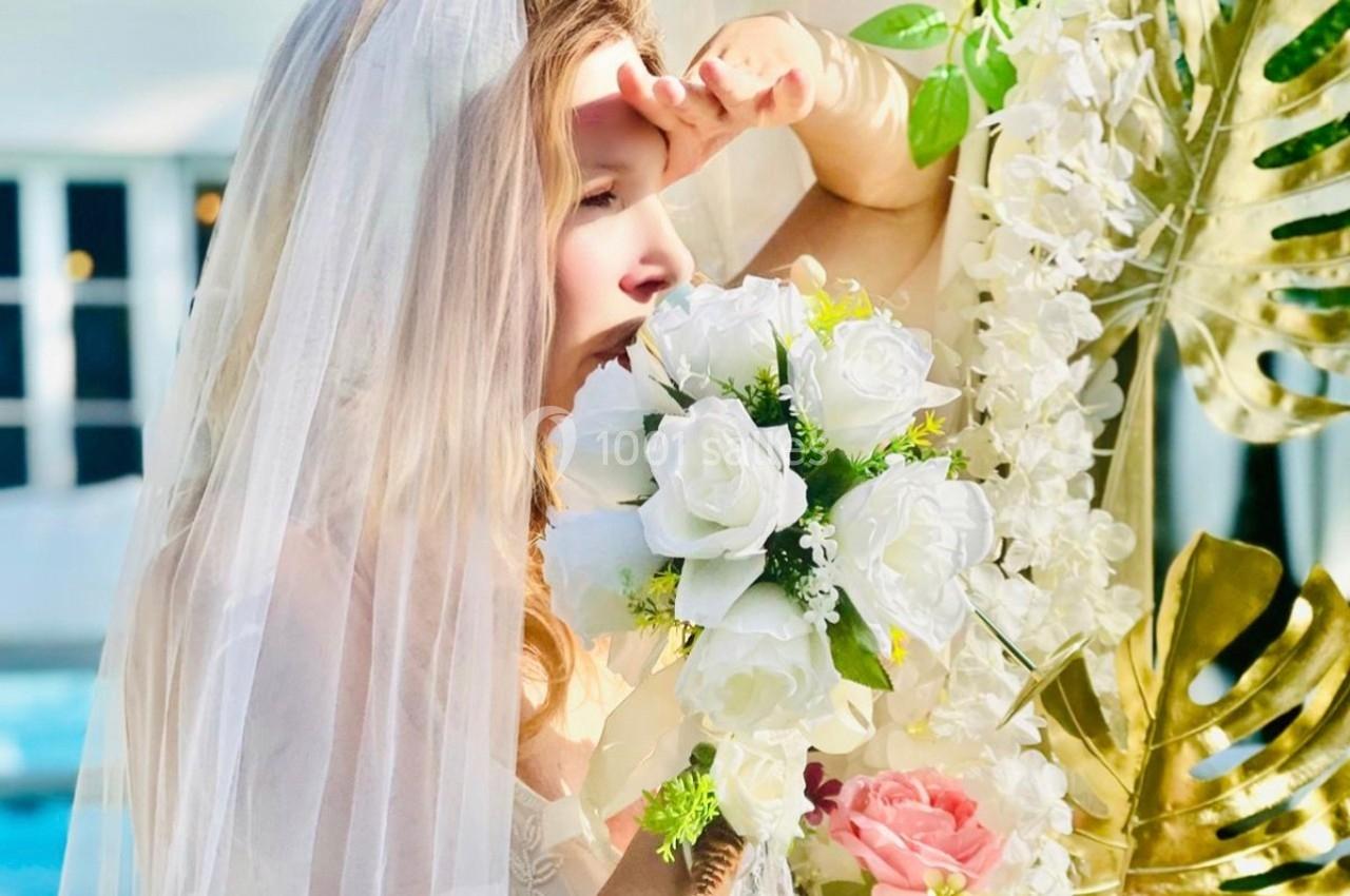 Femme en robe de mariée tenant un bouquet de fleurs blanches et roses près d'une arche décorée de feuillages.