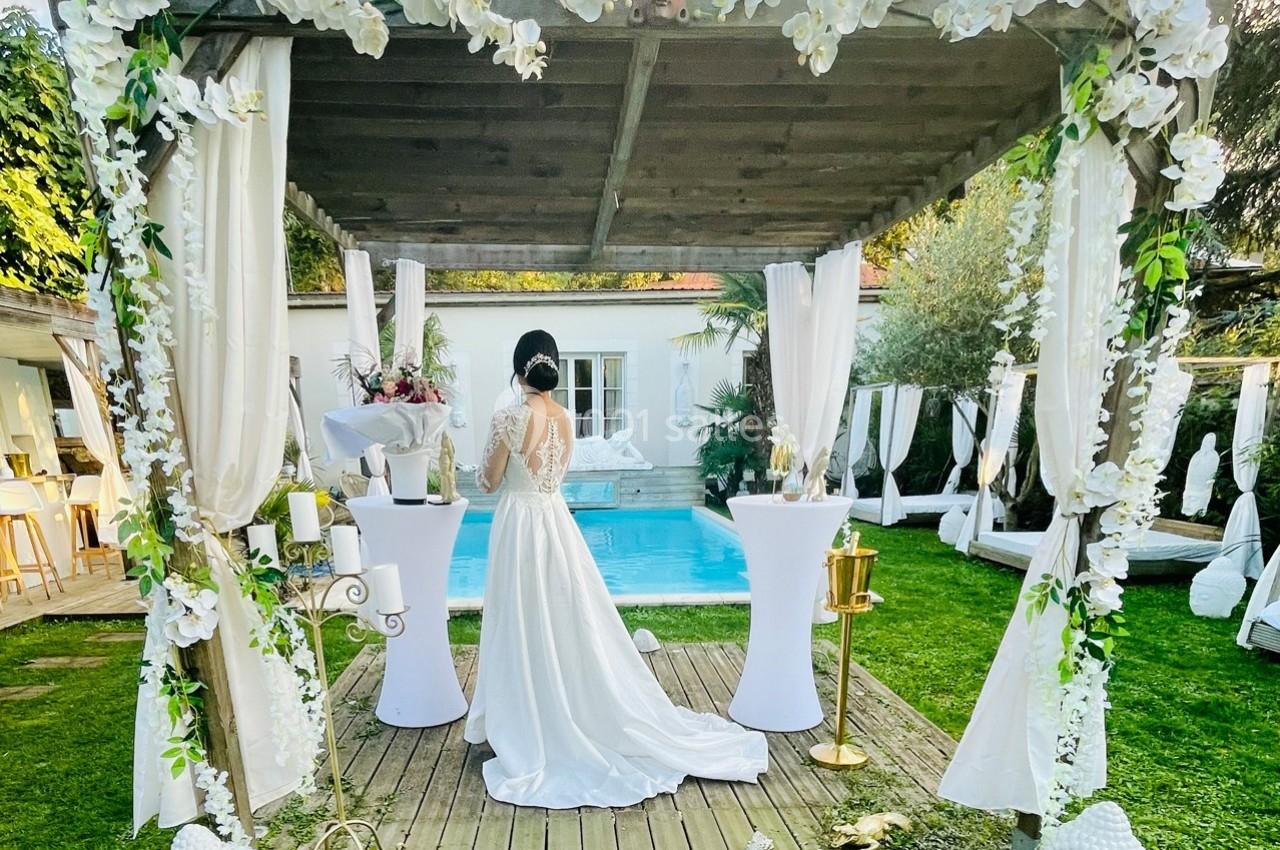 Une mariée en robe blanche se tient sous une pergola décorée, près d'une piscine dans un jardin.