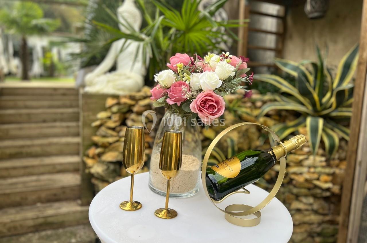 Vase de fleurs roses et blanches, flûtes dorées et bouteille de champagne sur une table blanche en extérieur.