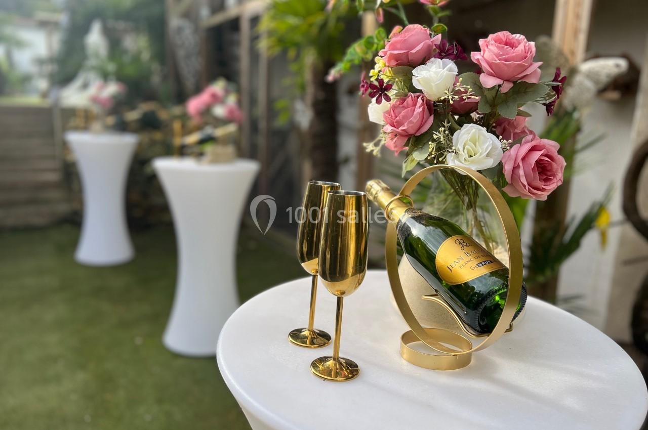 Bouteille de champagne avec deux flûtes dorées sur une table blanche, décorée de fleurs roses et blanches.