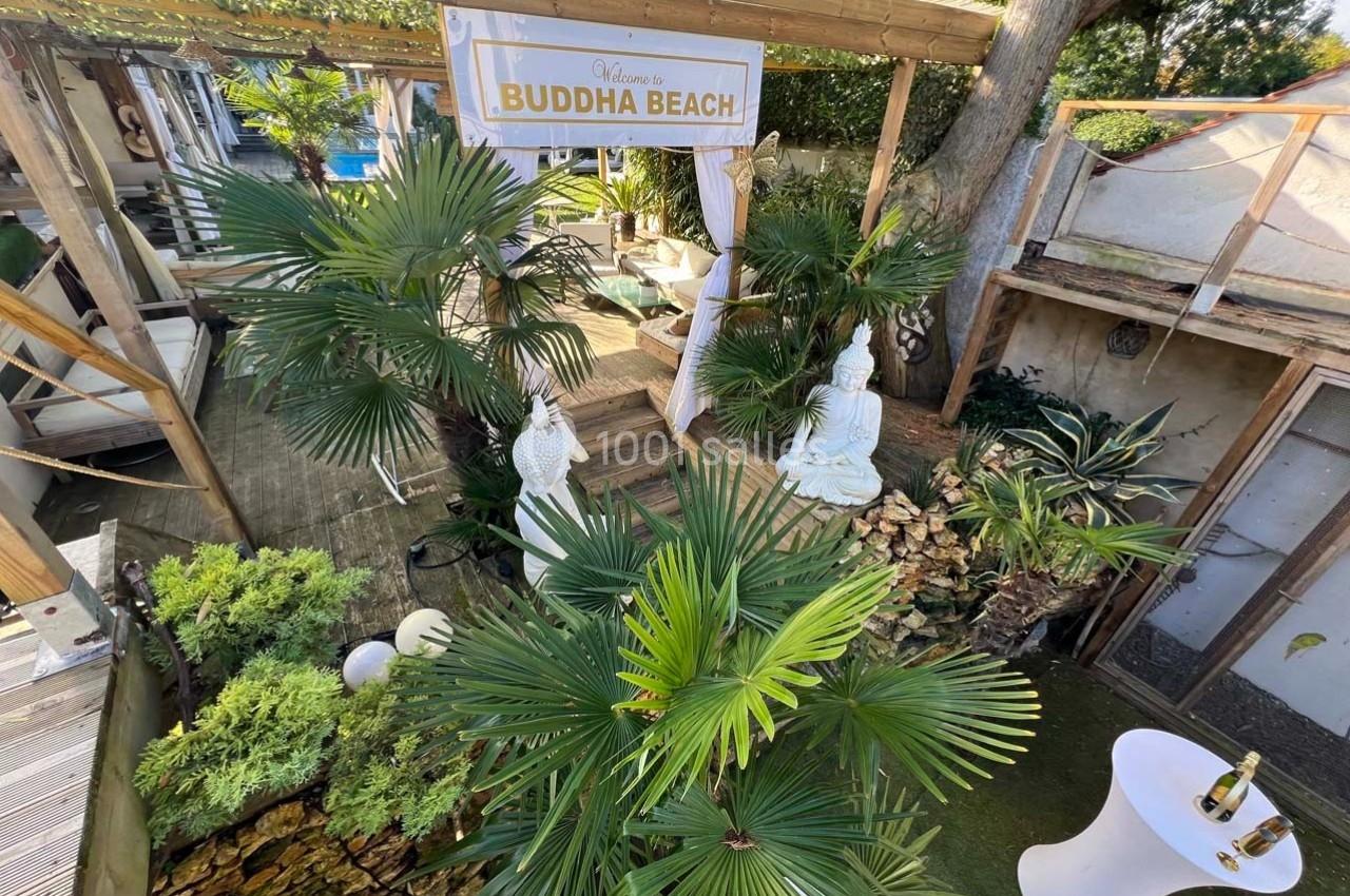 Jardin exotique avec statues de Bouddha, palmiers et terrasse en bois sous une pergola, ambiance paisible.