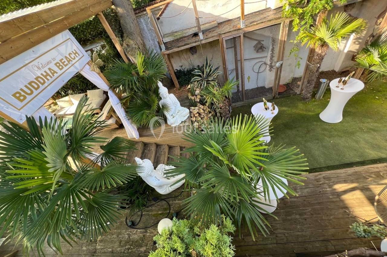 Vue d'une terrasse en bois avec des plantes tropicales, des statues blanches et des tables hautes dans un cadre extérieur.