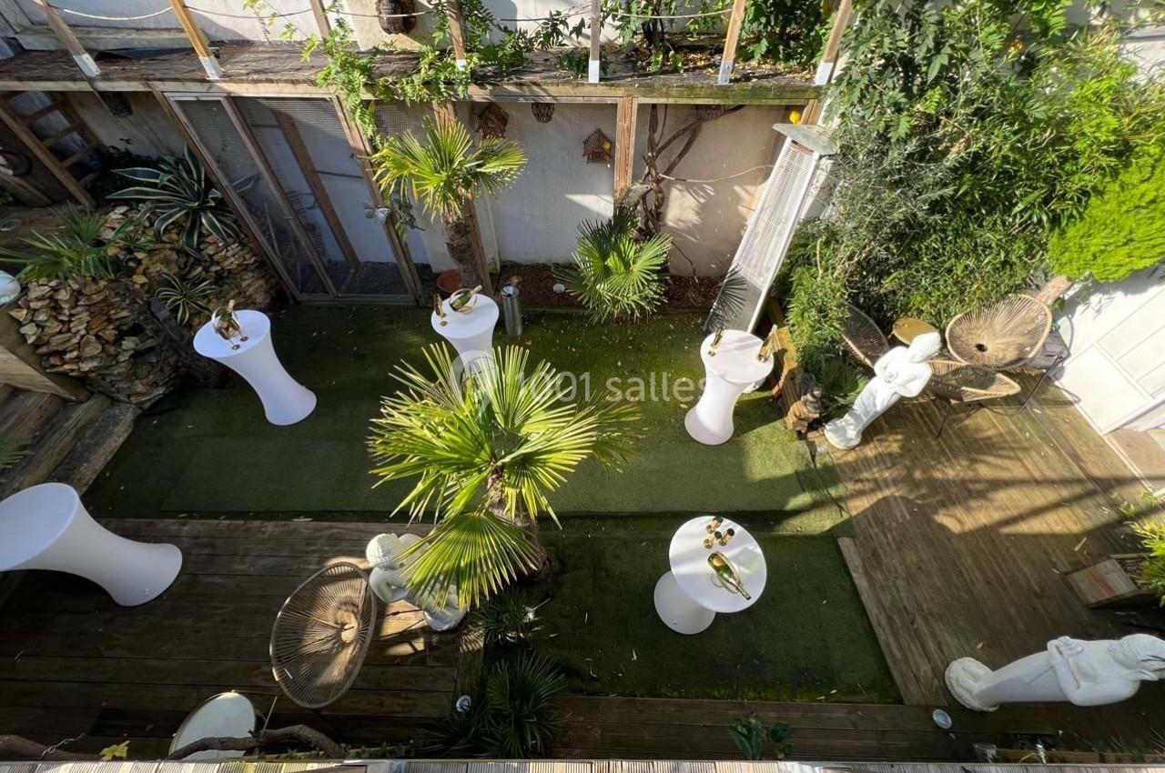 Vue d'une terrasse extérieure aménagée avec des plantes, des statues, des tables hautes et un sol en bois et gazon.