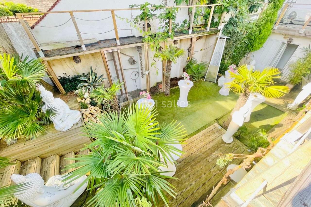Jardin aménagé avec statues, plantes exotiques, pelouse artificielle et terrasse en bois, entouré de murs blancs.