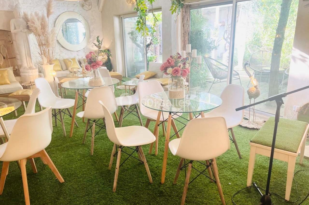 Salle lumineuse avec tables en verre, chaises blanches, décor floral et sol en gazon synthétique près de baies vitrées.