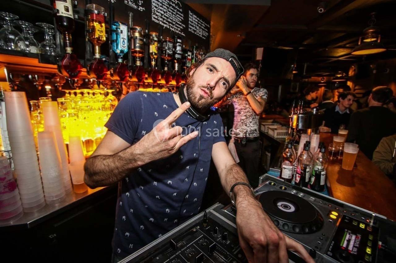 Un DJ portant une casquette à l'envers fait un geste de la main devant une console dans un bar éclairé.