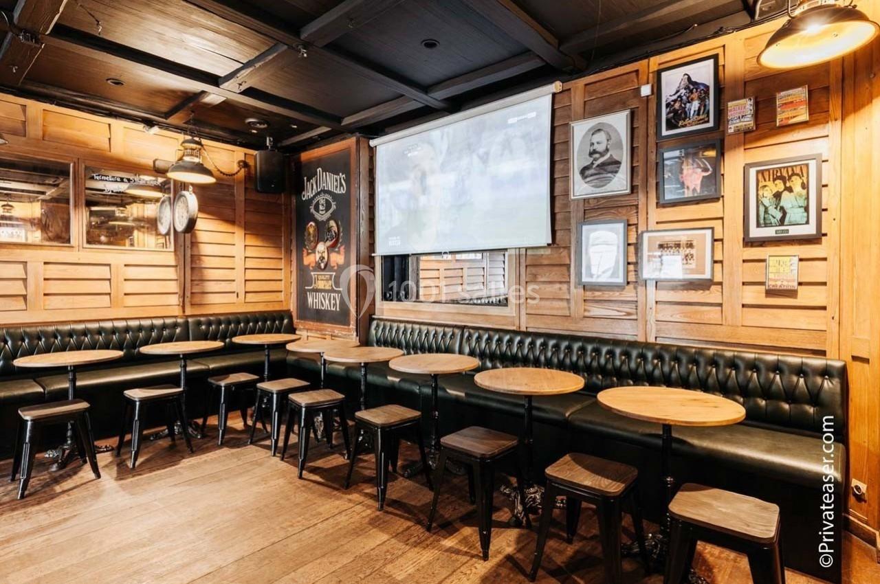 Intérieur d'un bar avec des tables rondes, des tabourets en bois et des murs décorés de cadres et affiches.