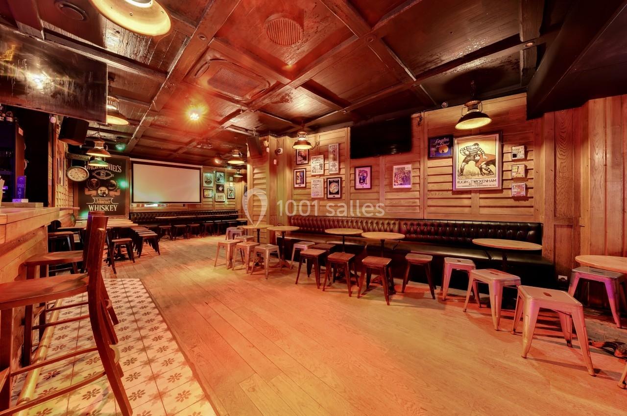 Intérieur d'un bar chaleureux avec des tables en bois, des tabourets, un écran mural et une décoration rétro.