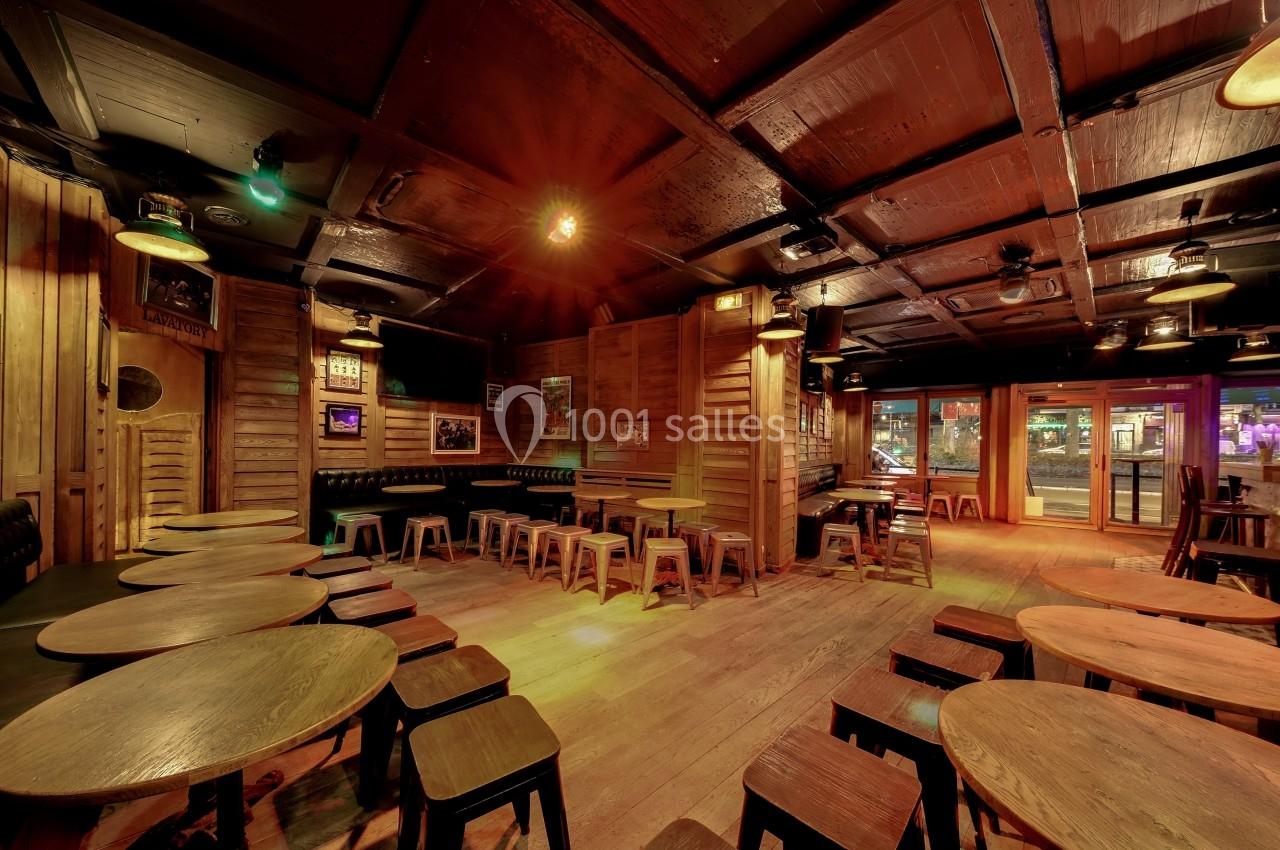 Salle de bar en bois avec tables rondes, tabourets et lumière tamisée, donnant sur une rue éclairée.