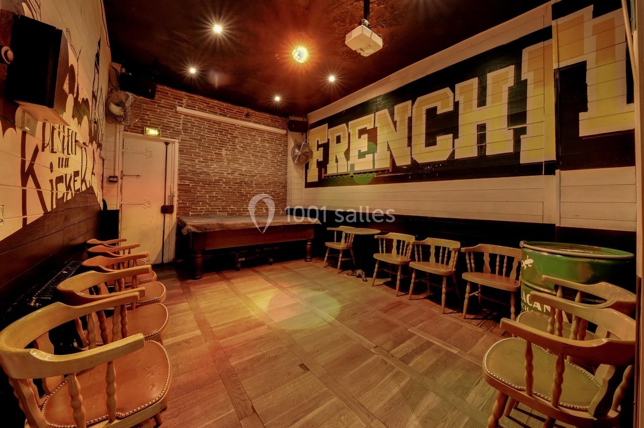 Salle vide avec des chaises en bois disposées autour de tables, murs en briques et peinture murale ’French’ en arrière-plan.