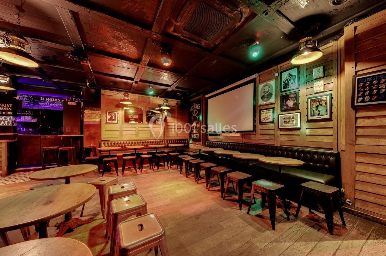 Salle de bar chaleureuse avec tables en bois, tabourets métalliques et écran de projection au mur.