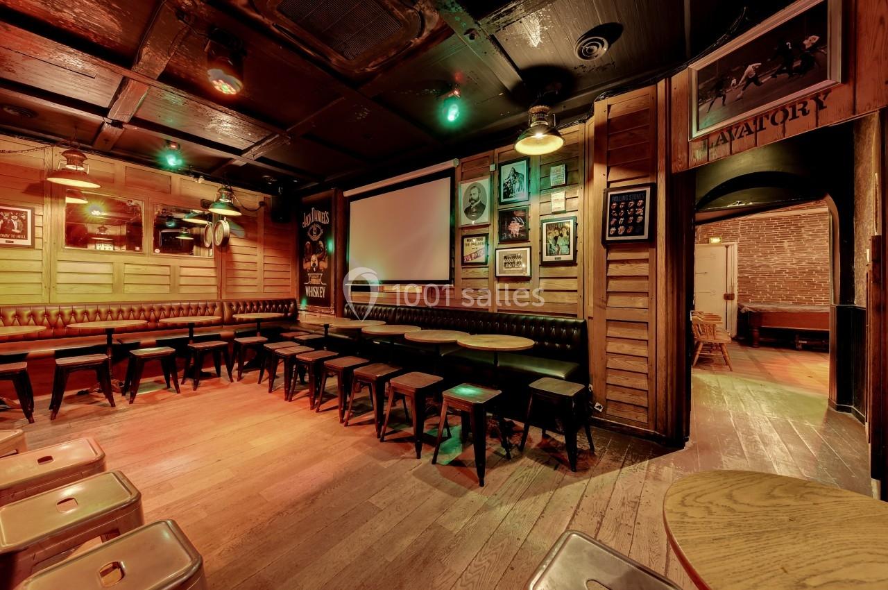 Salle de bar en bois avec tables, tabourets, écran mural et décorations vintage sur les murs.