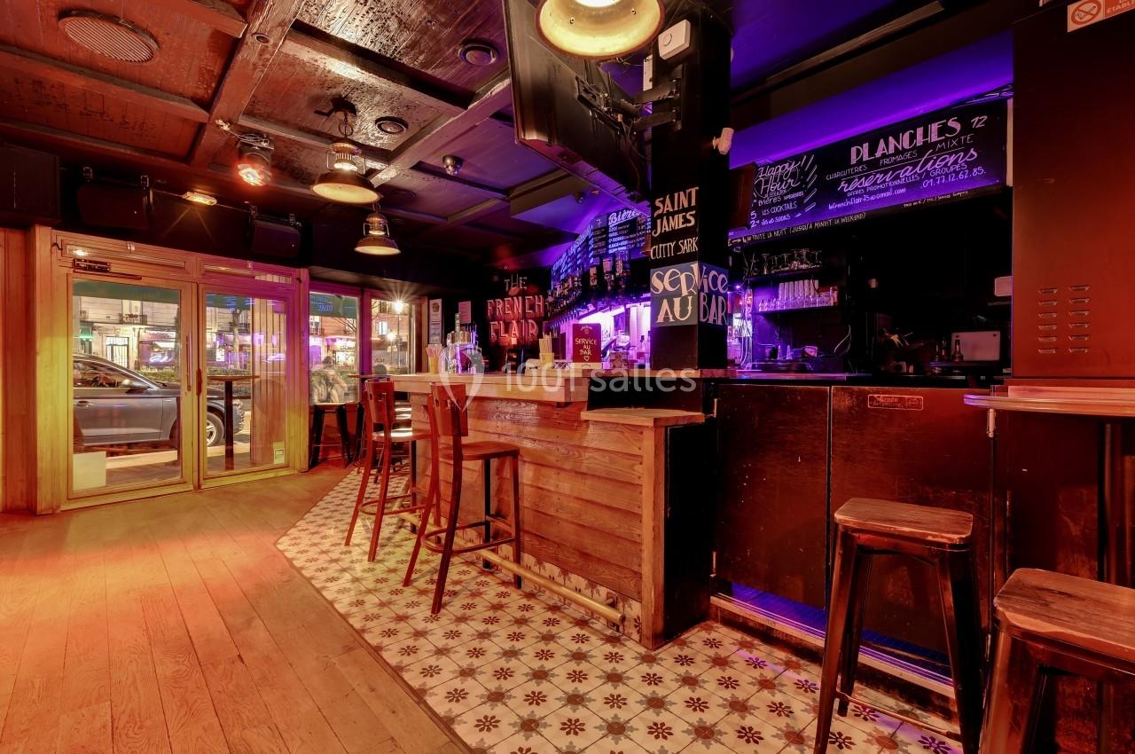 Intérieur d'un bar avec comptoir en bois, tabourets hauts, éclairage chaleureux et tableau noir affichant le menu.