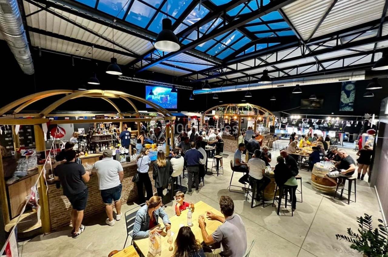 Salle animée avec des tables en bois, des stands de restauration et des groupes de personnes discutant sous un toit vitré.
