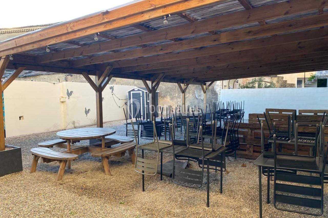 Terrasse couverte avec tables et chaises empilées sur un sol en gravier, entourée de murs peints et éclairée par des…