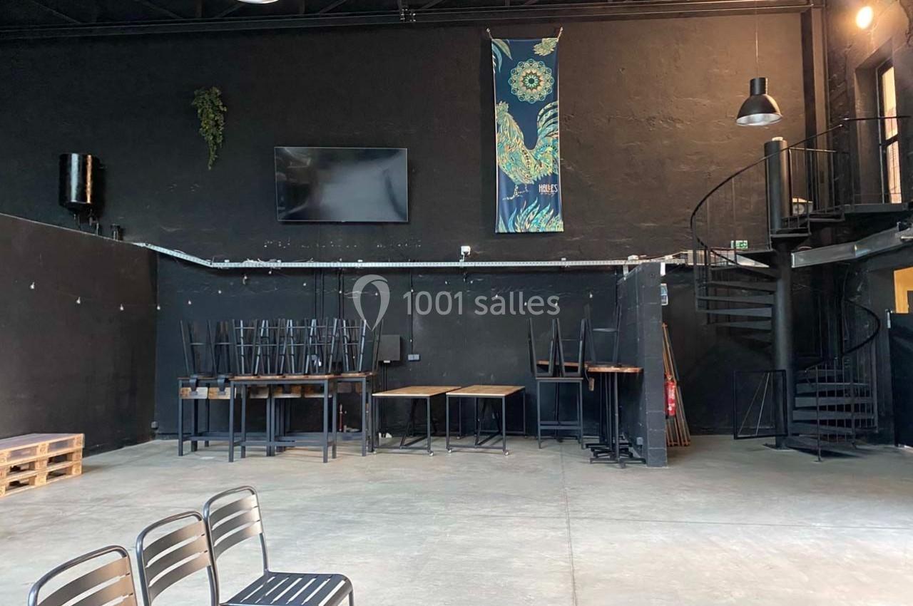 Salle vide avec murs noirs, tables empilées, chaises alignées, escalier en colimaçon et décoration murale colorée.
