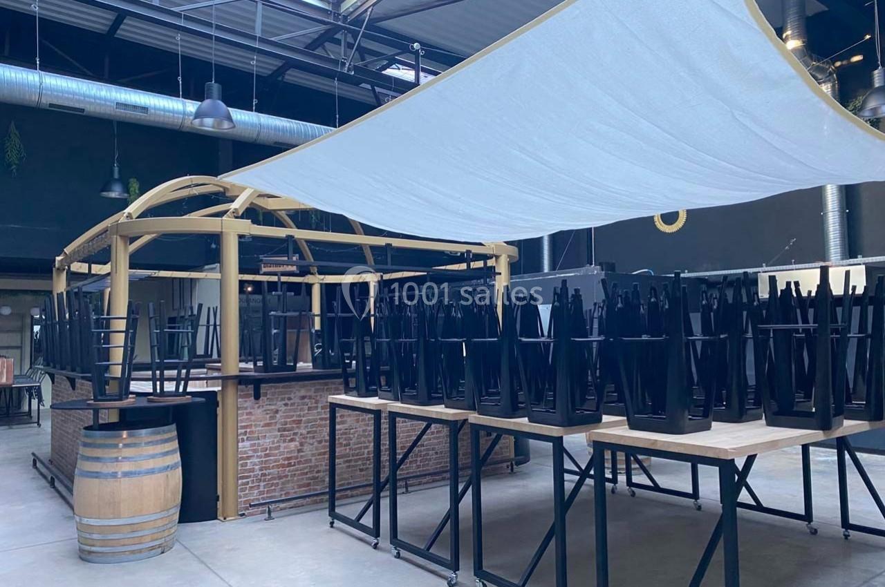 Chaises empilées sur des tables dans un espace intérieur avec un bar en briques et une grande toile tendue au plafond.