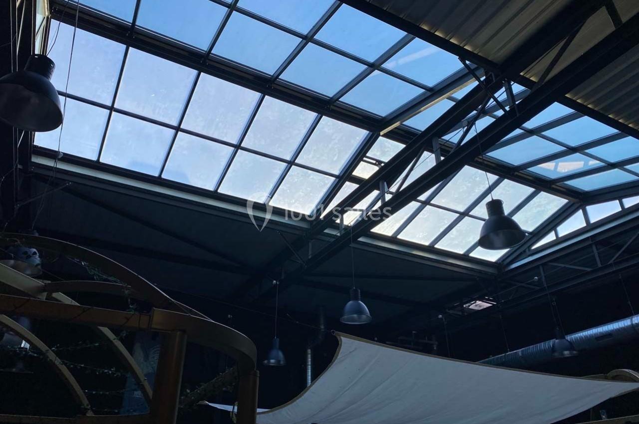 Plafond industriel avec grandes verrières laissant passer la lumière naturelle, éclairage suspendu visible.