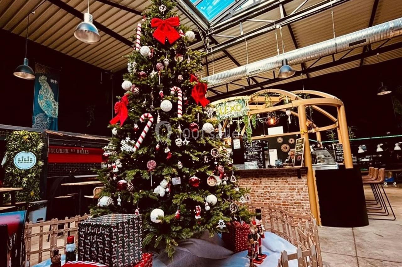 Sapin de Noël décoré avec guirlandes, boules et cadeaux à sa base, dans un espace intérieur au style industriel.