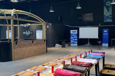 Salle aménagée avec des tables alignées portant des brochures, des bandanas colorés et des bougies, écran et affiches en…