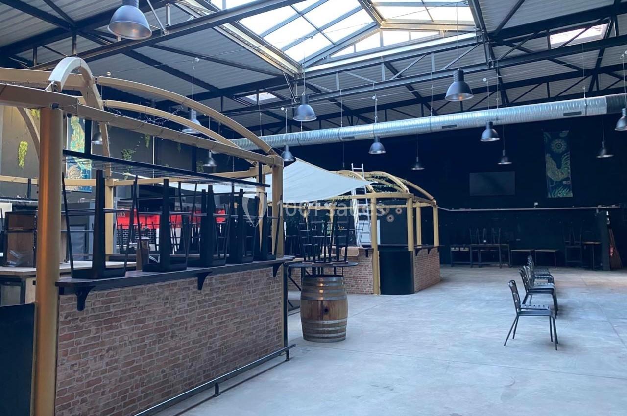 Intérieur d'un grand espace industriel avec des tables et chaises empilées, éclairé par une verrière.