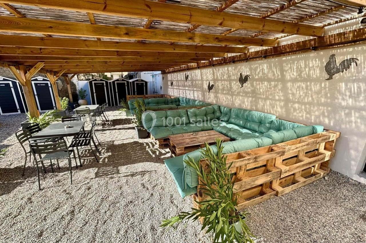 Terrasse ombragée avec salon en palettes et coussins verts, tables en métal noir, sol en gravier et décor mural.