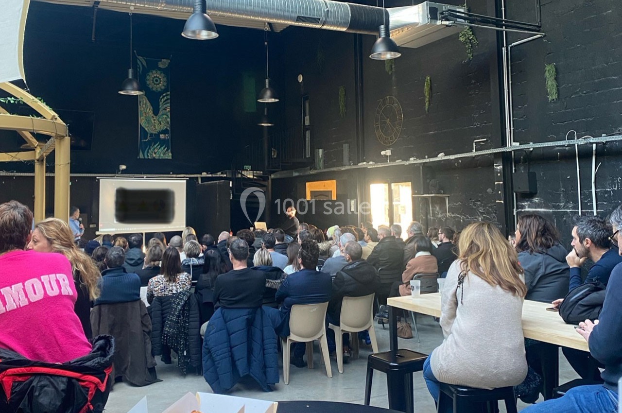 Un groupe de personnes assiste à une présentation dans une grande salle sombre avec des tables et des chaises.