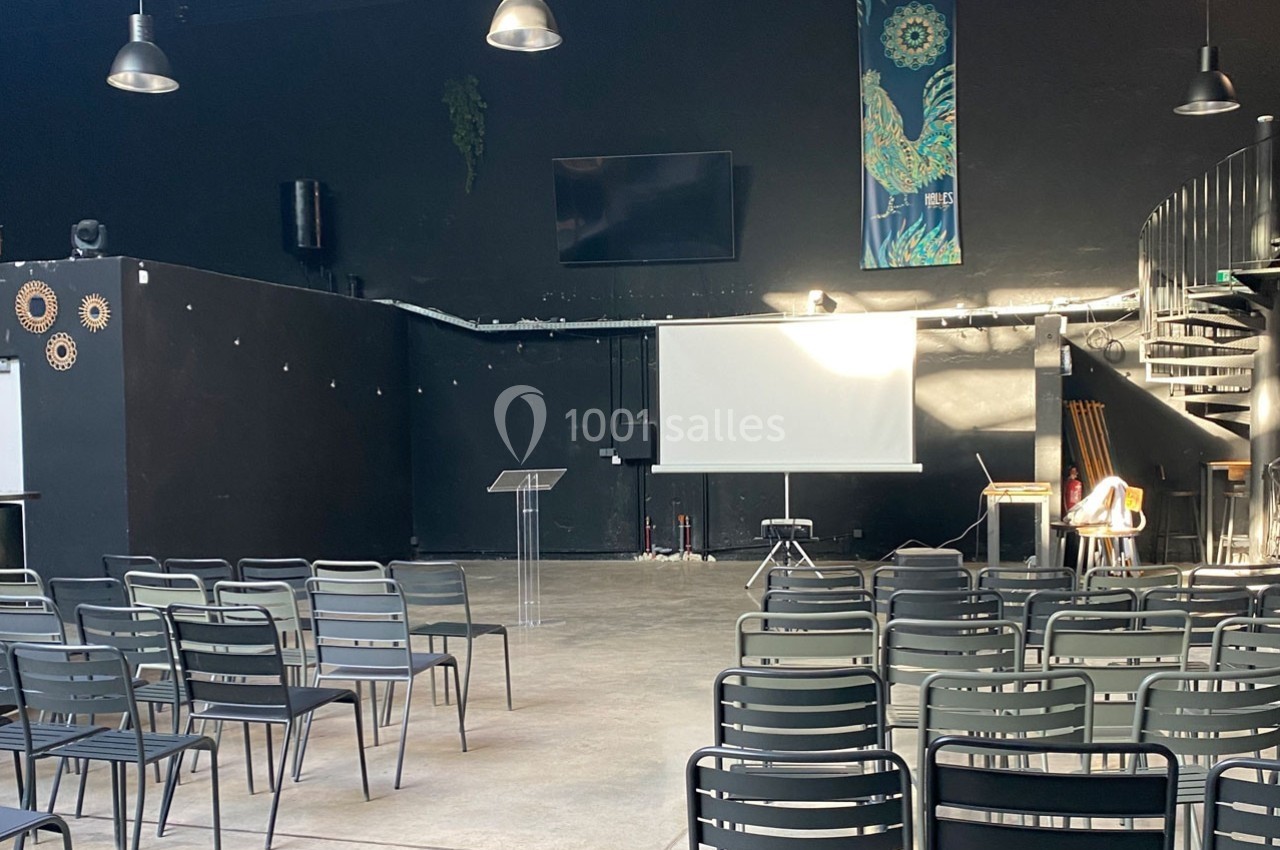 Salle sombre avec des chaises alignées, un écran de projection, un pupitre transparent et un escalier en colimaçon.