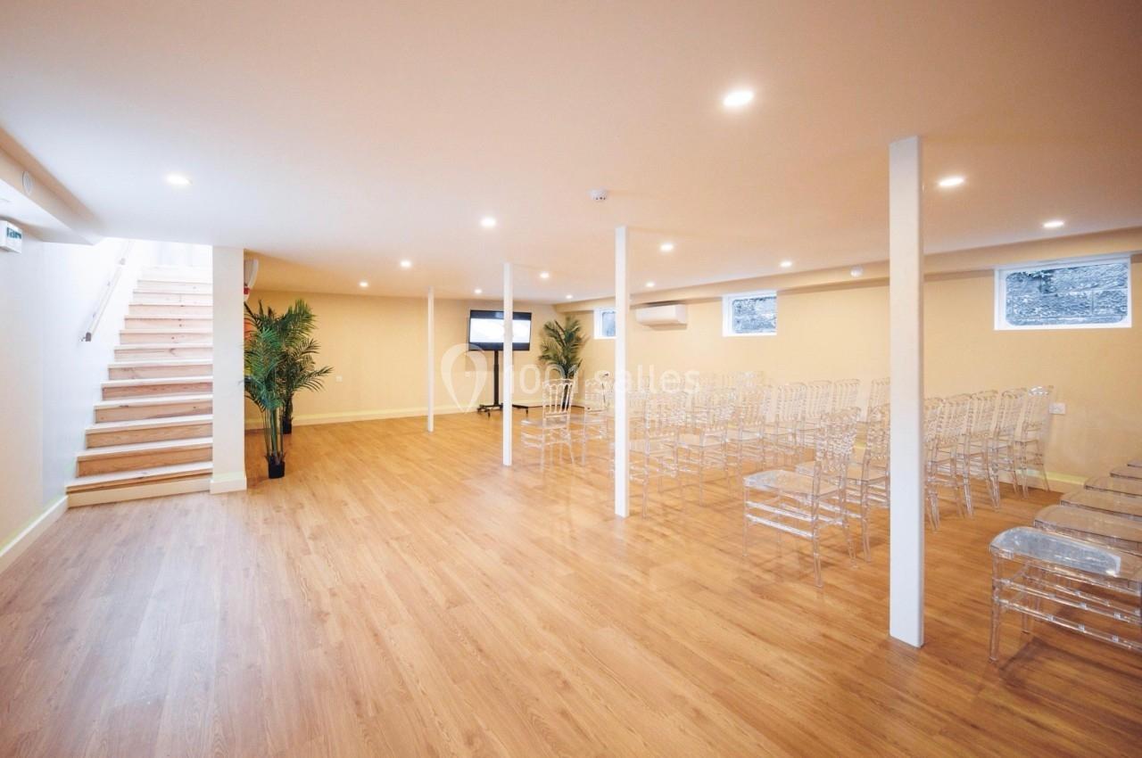 Salle lumineuse avec parquet, chaises transparentes alignées, écran au fond et escalier sur la gauche.