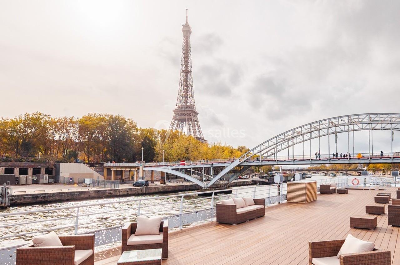 Vue sur la Tour Eiffel depuis une terrasse en bois au bord de la Seine, avec un pont et des arbres en arrière-plan.