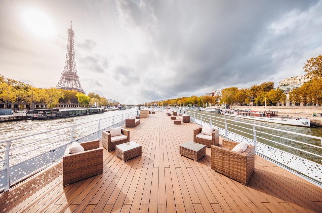 Pont d'un bateau avec des fauteuils en rotin, vue sur la Seine et la tour Eiffel sous un ciel nuageux.