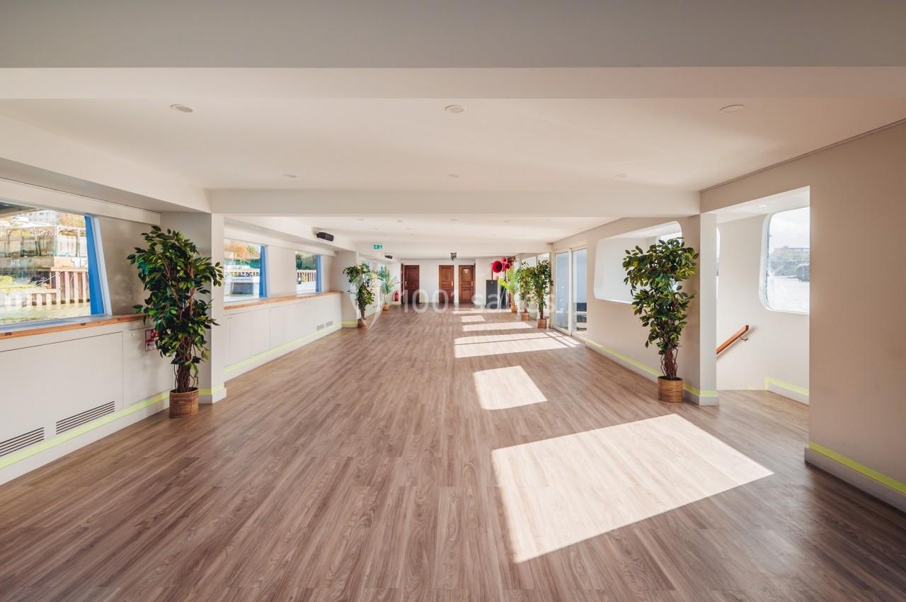 Salle lumineuse avec parquet, grandes fenêtres laissant entrer la lumière naturelle, et plantes en pot alignées.