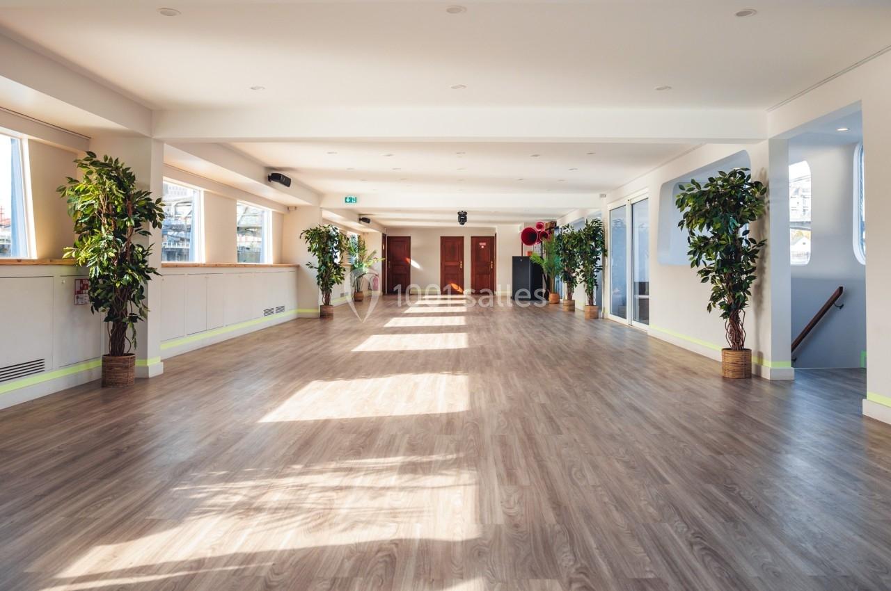 Salle lumineuse avec parquet en bois, grandes fenêtres laissant entrer la lumière et plantes disposées le long des murs.