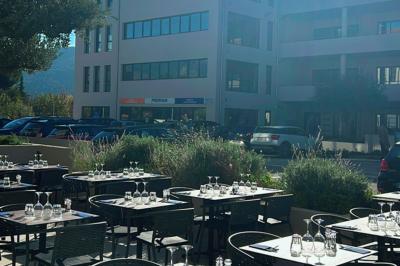 Terrasse d'un restaurant avec des tables dressées, située devant un immeuble moderne et un parking ensoleillé.