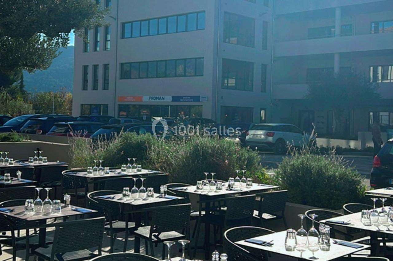 Terrasse d'un restaurant avec des tables dressées, située devant un immeuble moderne et un parking ensoleillé.