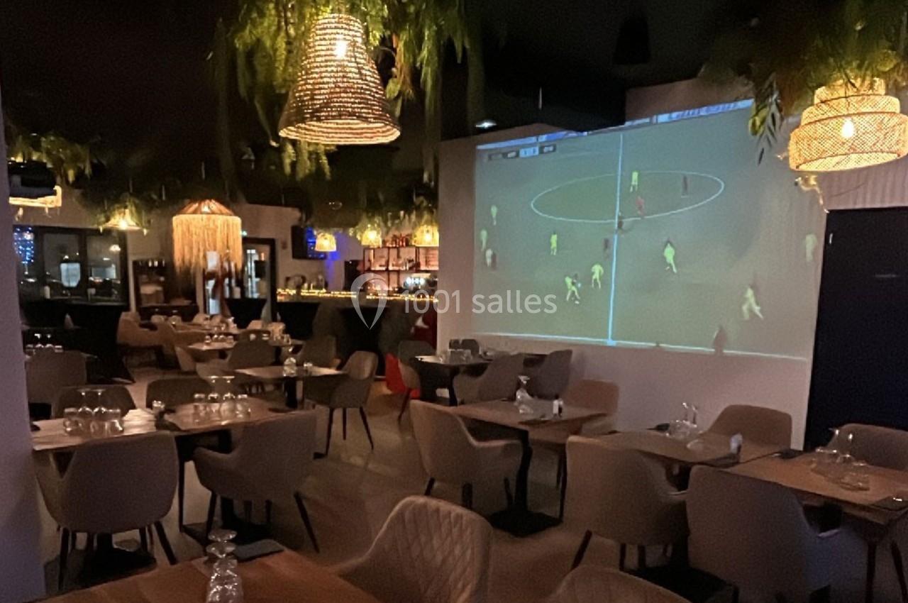 Salle de restaurant avec tables dressées, éclairage tamisé et un écran diffusant un match de football.