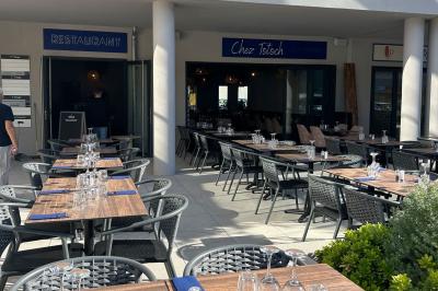 Terrasse d'un restaurant avec des tables dressées, située devant un immeuble moderne et un parking ensoleillé.