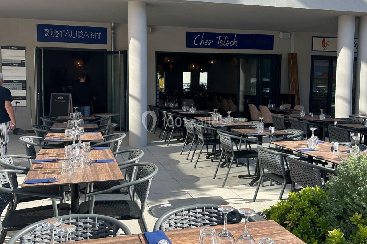 Terrasse d'un restaurant avec tables dressées, chaises en osier et entrée visible sous un auvent.
