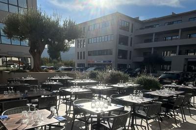 Terrasse d'un restaurant avec des tables dressées, située devant un immeuble moderne et un parking ensoleillé.