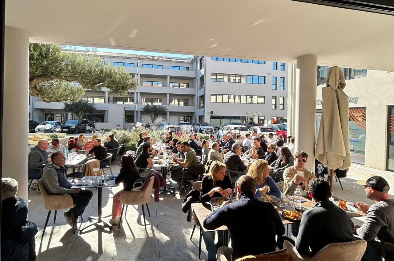 Des personnes assises en terrasse d'un restaurant, partageant un repas en plein jour, entourées de bâtiments modernes.