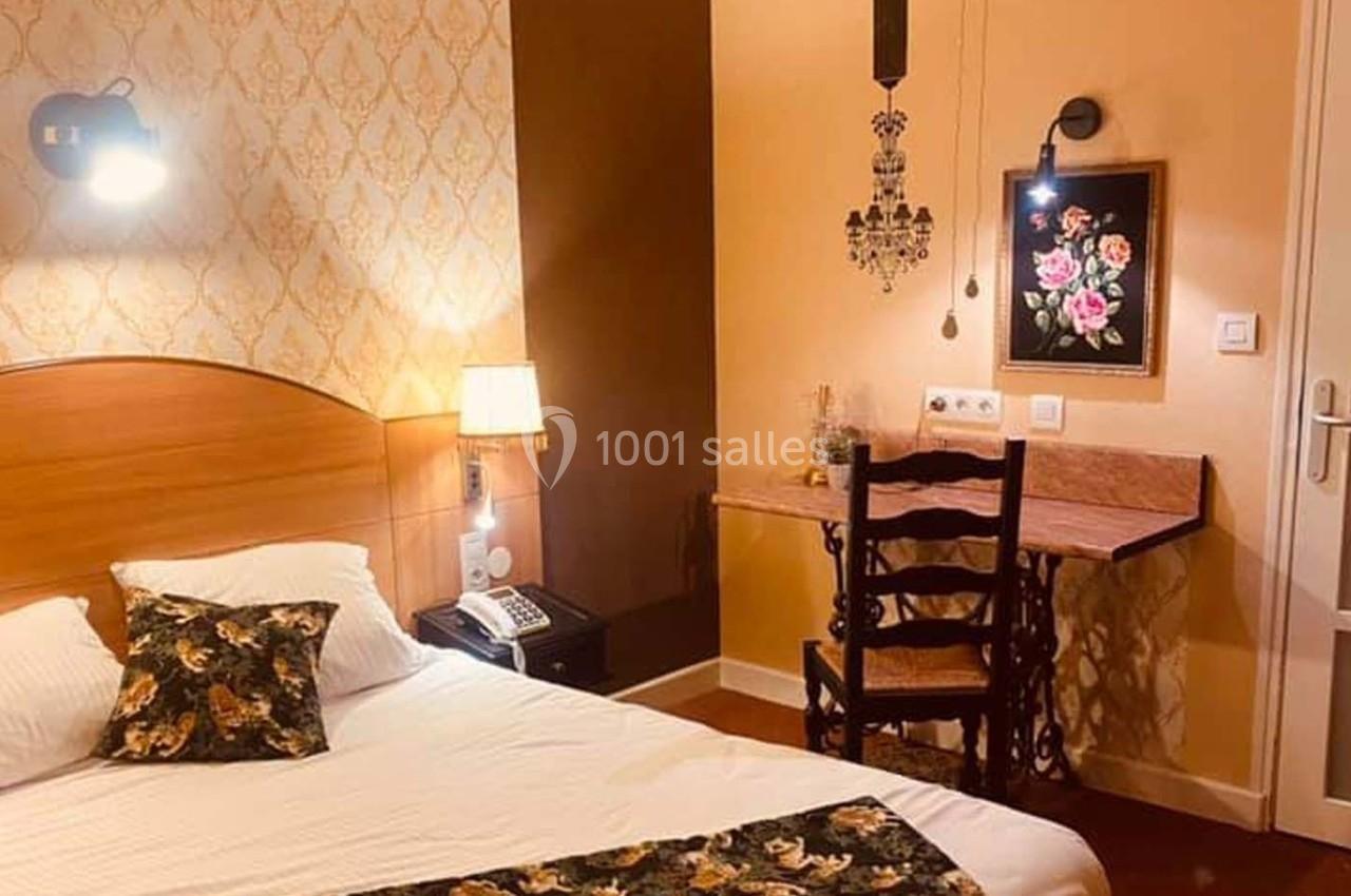 Chambre avec lit double, bureau en bois, chaise, lampe murale et décoration florale sur un mur beige et brun.