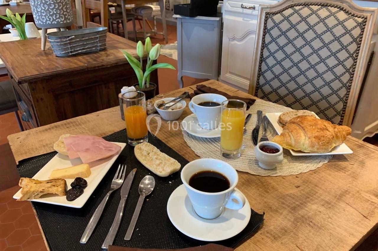 Petit-déjeuner servi sur une table en bois avec café, jus d'orange, croissant, pain, confiture, jambon et décor végétal.