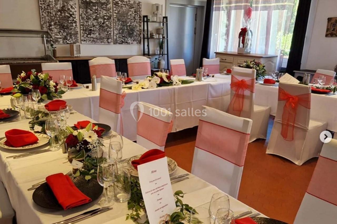 Salle de réception décorée avec des tables dressées, nappes blanches, serviettes rouges et chaises ornées de rubans roses.