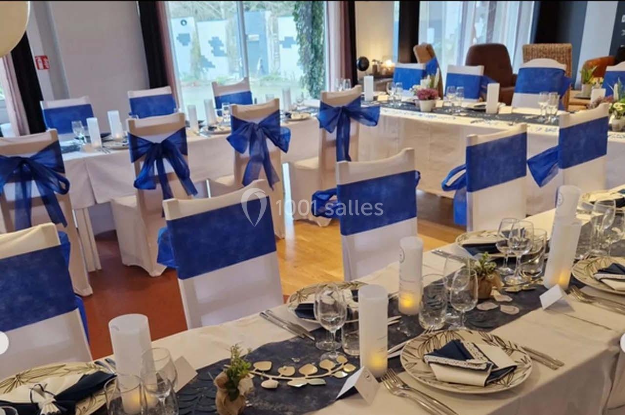 Salle de réception décorée avec des nappes blanches, des chaises ornées de nœuds bleus et des tables dressées élégamment.