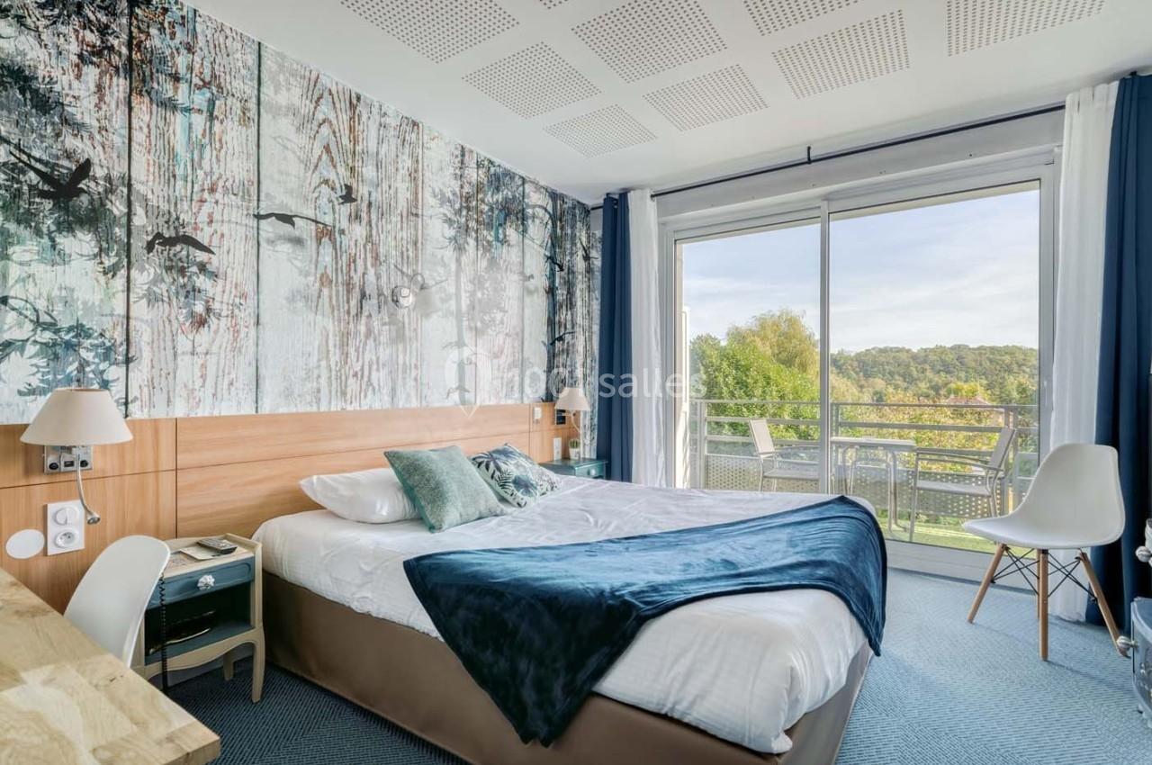 Chambre lumineuse avec lit double, décoration murale nature, bureau, et balcon donnant sur un paysage verdoyant.