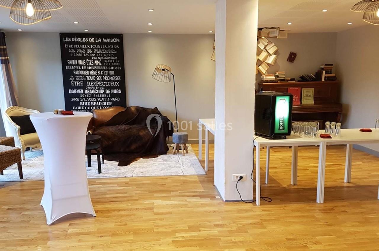 Salle lumineuse avec parquet, coin salon, tables blanches, distributeur de boissons et piano en arrière-plan.