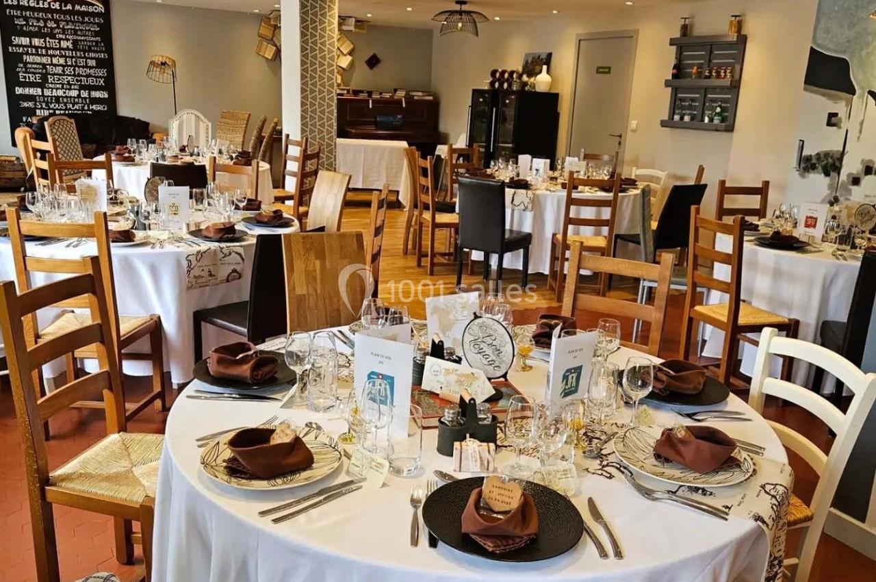 Salle de restaurant décorée avec des tables rondes dressées, nappes blanches, serviettes marron et chaises en bois.