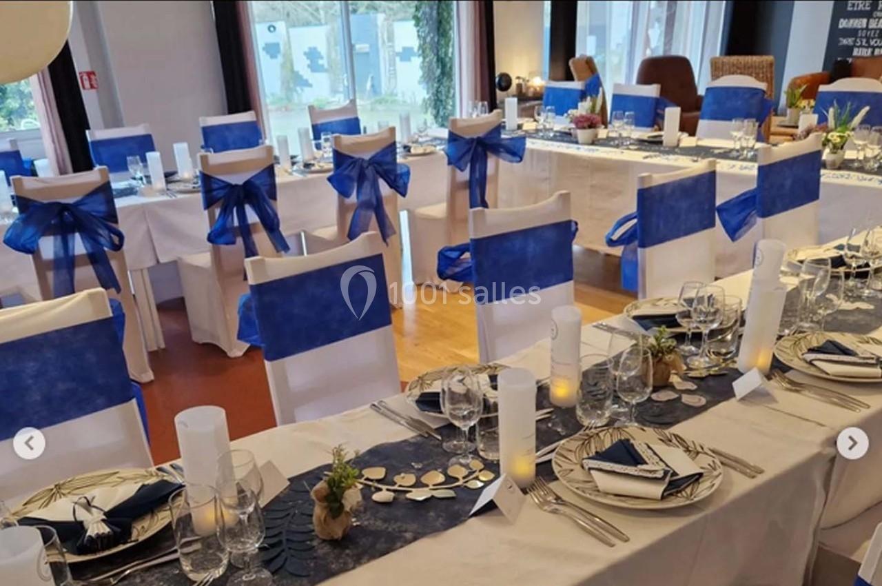 Salle de réception décorée avec des tables dressées, nappes blanches et accents bleus, chaises assorties avec nœuds bleus.